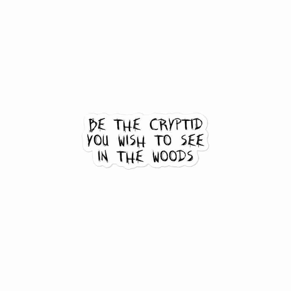 A sticker of uppercase text reading "Be the cryptid you wish to see in the woods".